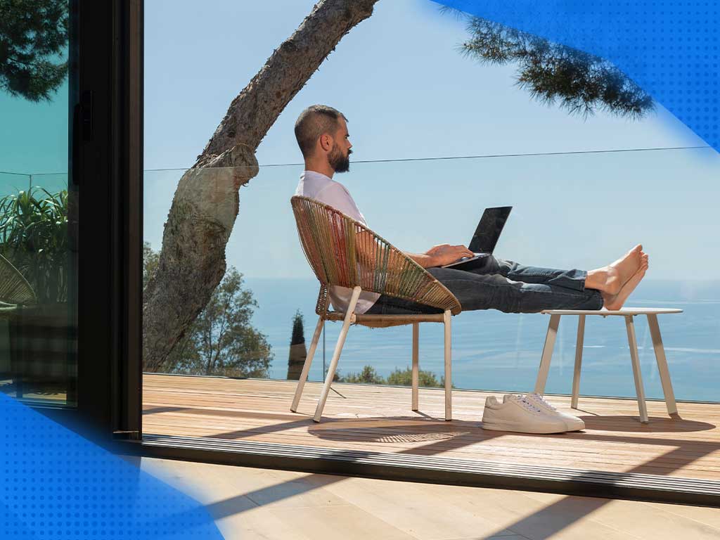 Mężczyzna siedzący na balkonie przy plaży. Mężczyzna ma laptop na nogach co wskazuje na pracę zdalną. Praca zdalna może być wykonywana z każdego miejsca na ziemi - jeśli ma się dostęp do Internetu. 